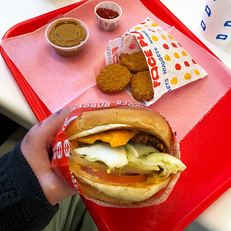 an order of chicken nuggests with dipping sauces on a tray with someone holding a burger in the forefront.