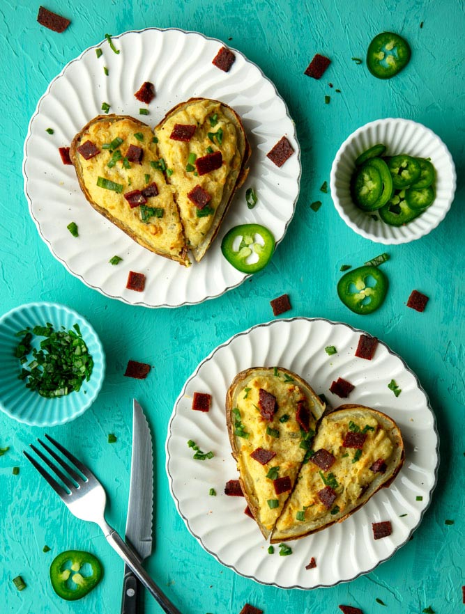 two heart shaped vegan twice baked potatoes, garnished with chives, bacon, and jalepeños